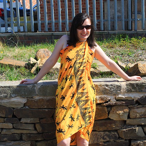 A woman wearing a yellow sarong patterned with black lizards leans against a low stone wall.