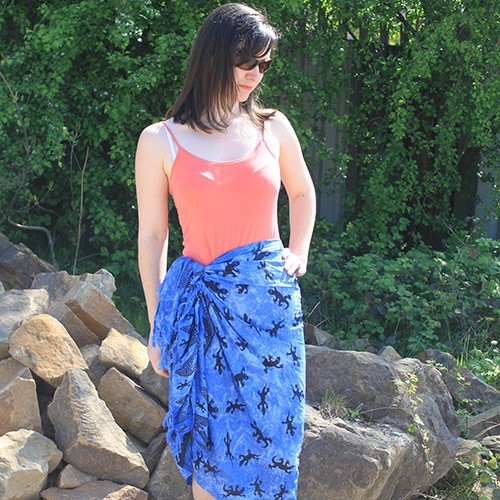 A woman ties a blue lizard-print sarong around her waist over a coral tank top, standing amidst a pile of rocks and lush greenery.