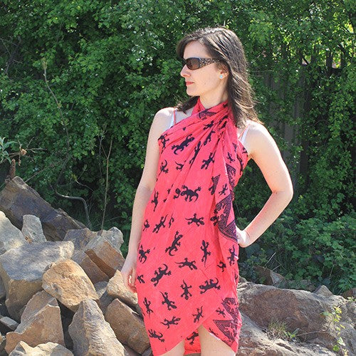 A woman wears a coral sarong patterned with black lizards. She stands outdoors amidst rocks and greenery.