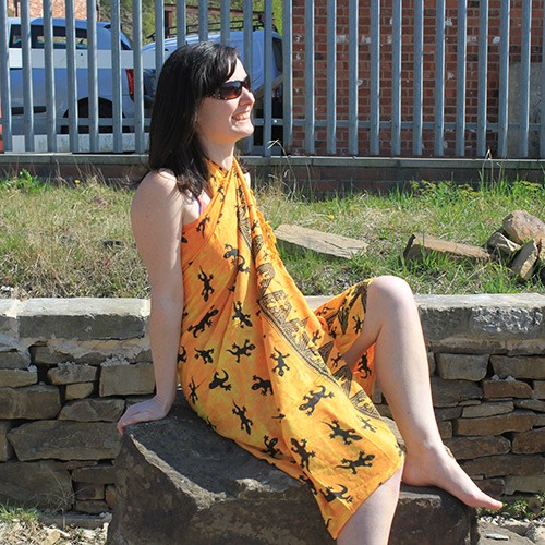 A woman sits on a rock, wearing a yellow sarong with a black lizard print, outdoors near a fence.
