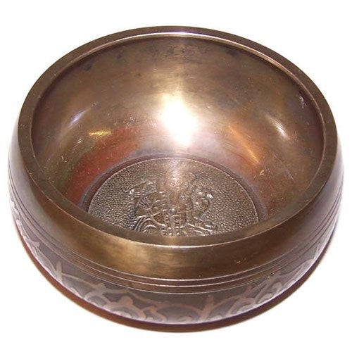 A bronze singing bowl with intricate engravings rests on a white surface, reflecting light. The interior has a detailed pattern at the center, adding to its ornate design.