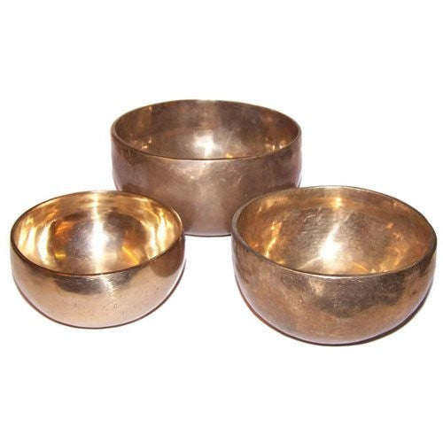 Three metallic singing bowls rest on a flat, white surface, with each bowl showcasing a polished, reflective finish. The bowls vary in size, sitting close together in a triangular arrangement.