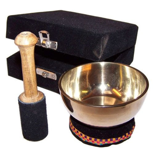 A metallic singing bowl and wooden mallet rest beside a decorative cushion. A black box with metal latches is in the background, indicating the items' storage container.