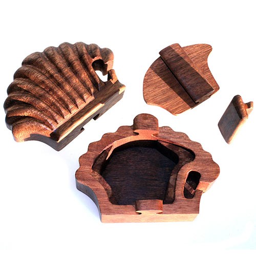 A carved wooden puzzle box lies open, revealing its compartment. The shell-shaped lid and interlocking pieces are separated, set against a plain white background. No text is present.