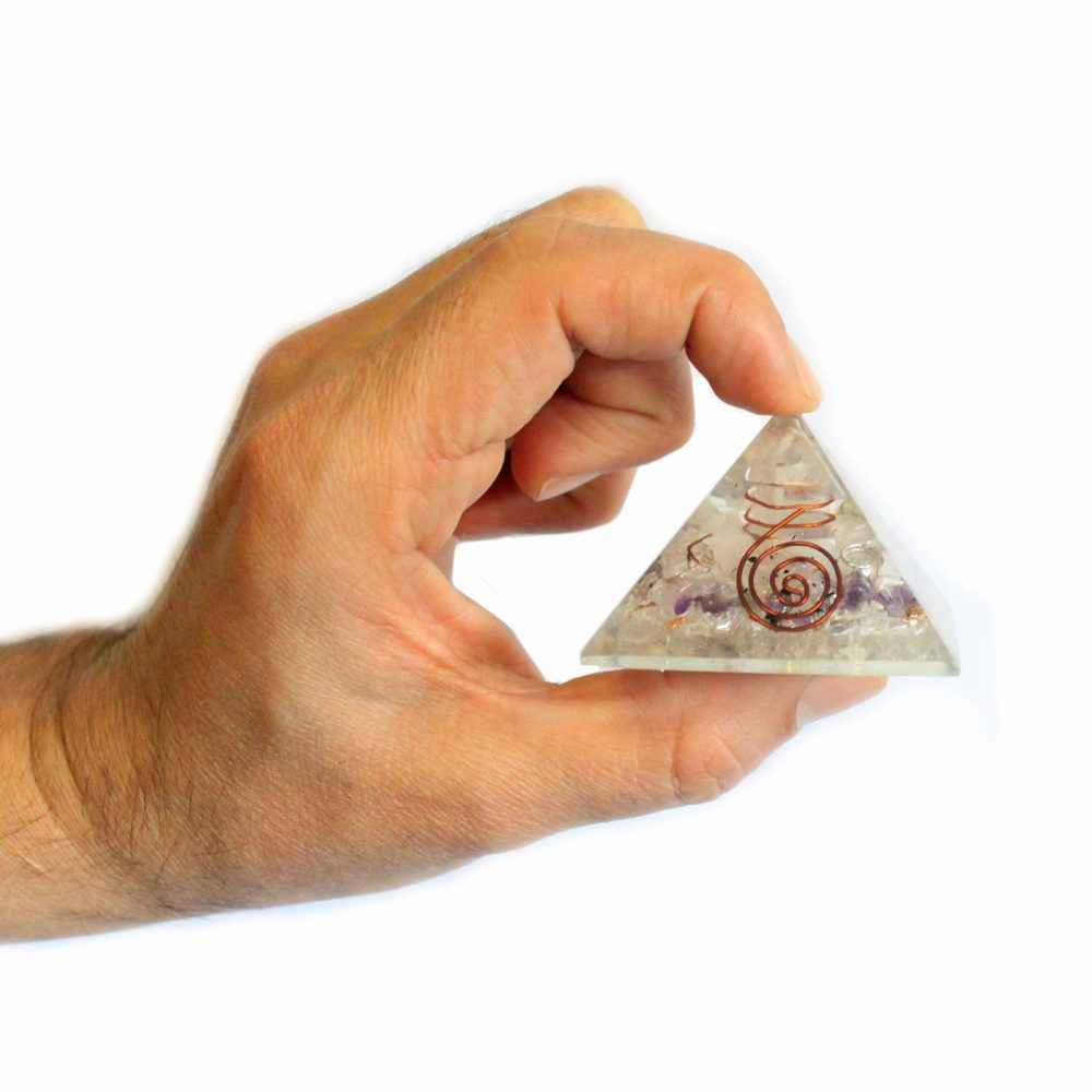 A hand holds a small, clear quartz pyramid containing amethyst and copper wire. The pyramid is embedded with small stones and a spiral of copper wire.