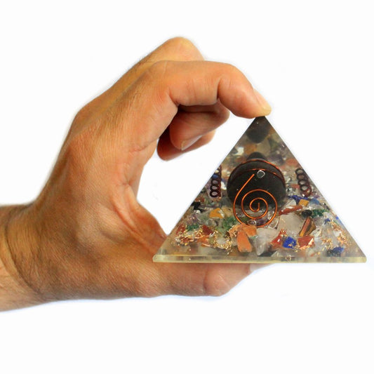 A hand holds a clear crystal pyramid embedded with various colored stones and a copper coil; the pyramid is small and rests in the palm.