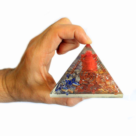 A hand holds a clear quartz pyramid containing embedded multicolored stones and a copper wire spiral. The pyramid is positioned upright. The background is white.