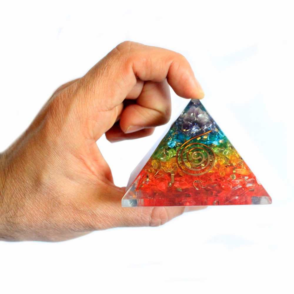 A hand is holding the apex of a crystal pyramid containing embedded spirals and red, orange, yellow, green, blue, and purple crystals against a white background.
