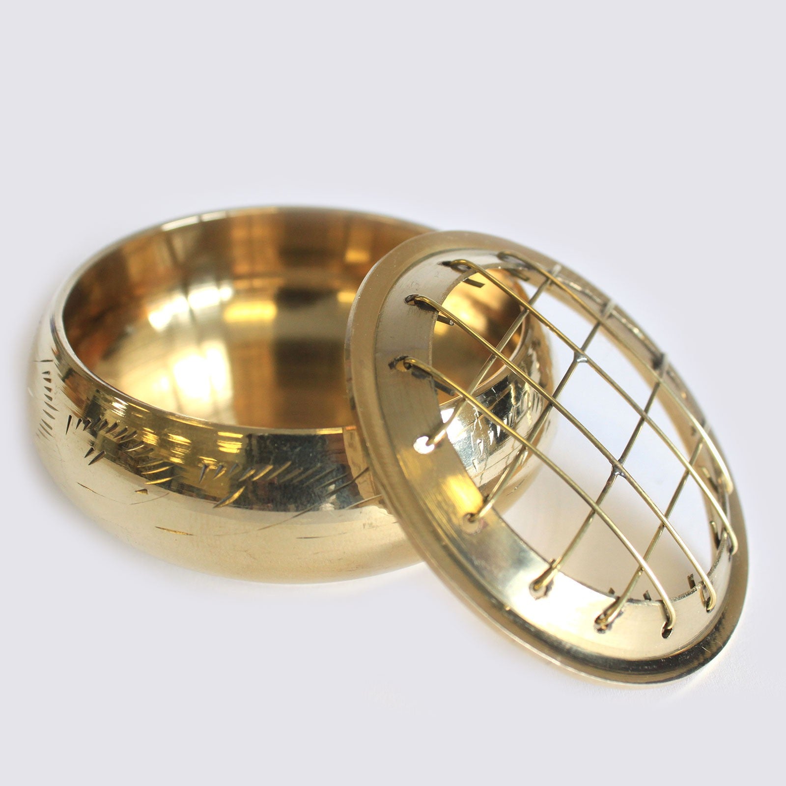A brass pot with engraving rests open, featuring a removable lid with a grid design, on a plain, light background.