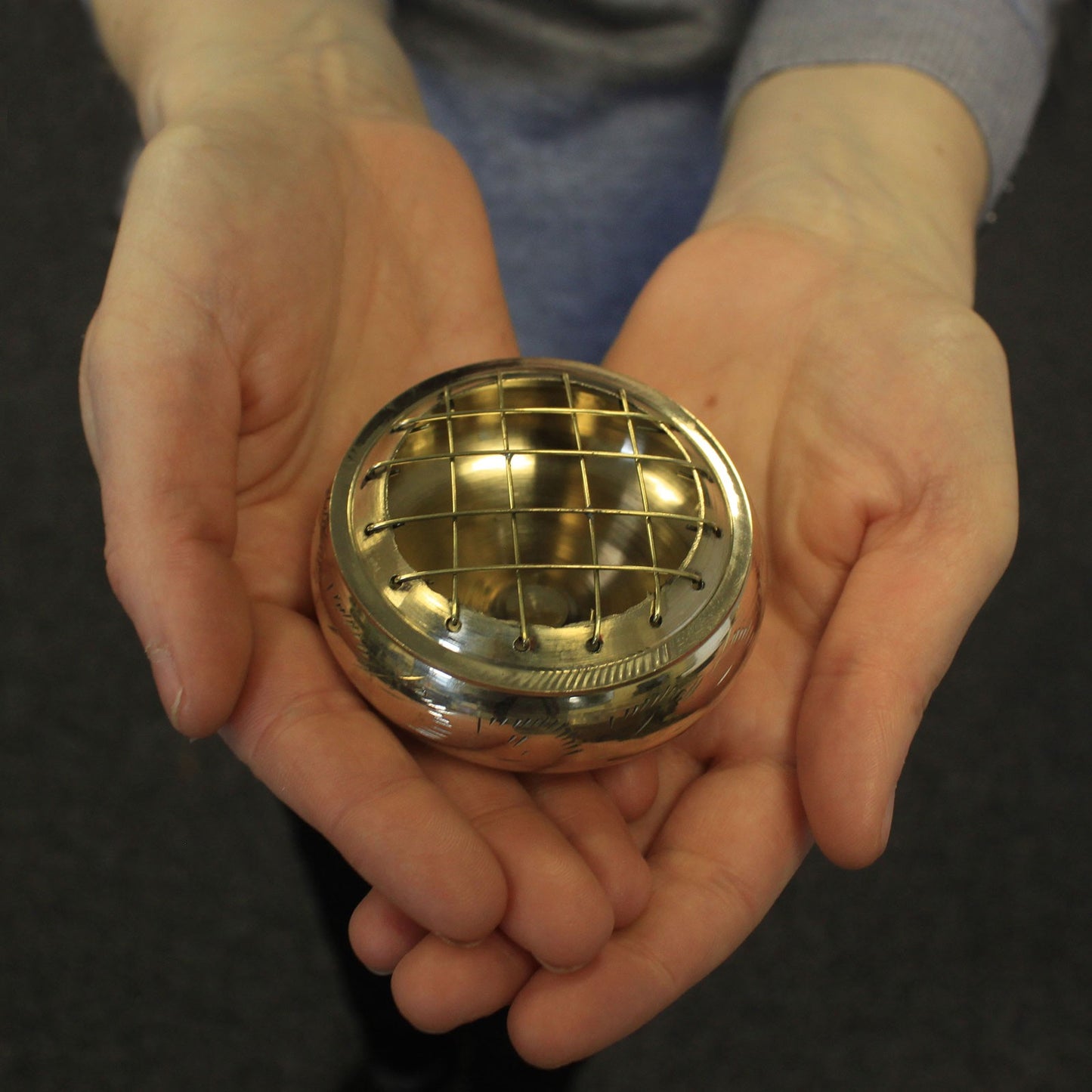 A metal object with a grid pattern rests in open hands. The textured, domed top reflects light. The person holds it gently in an indoor setting.