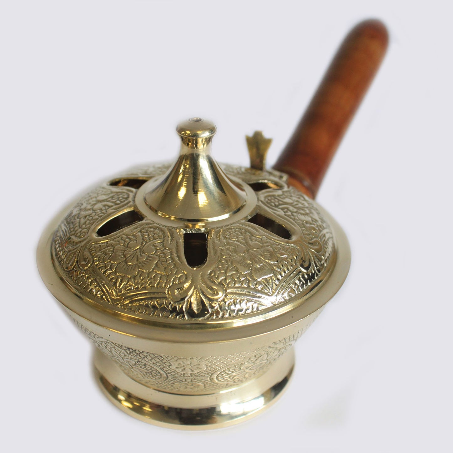 A brass incense burner with intricate floral engravings sits with a wooden handle extending outward, designed for holding and dispensing aromatic smoke. The background is a plain light surface.