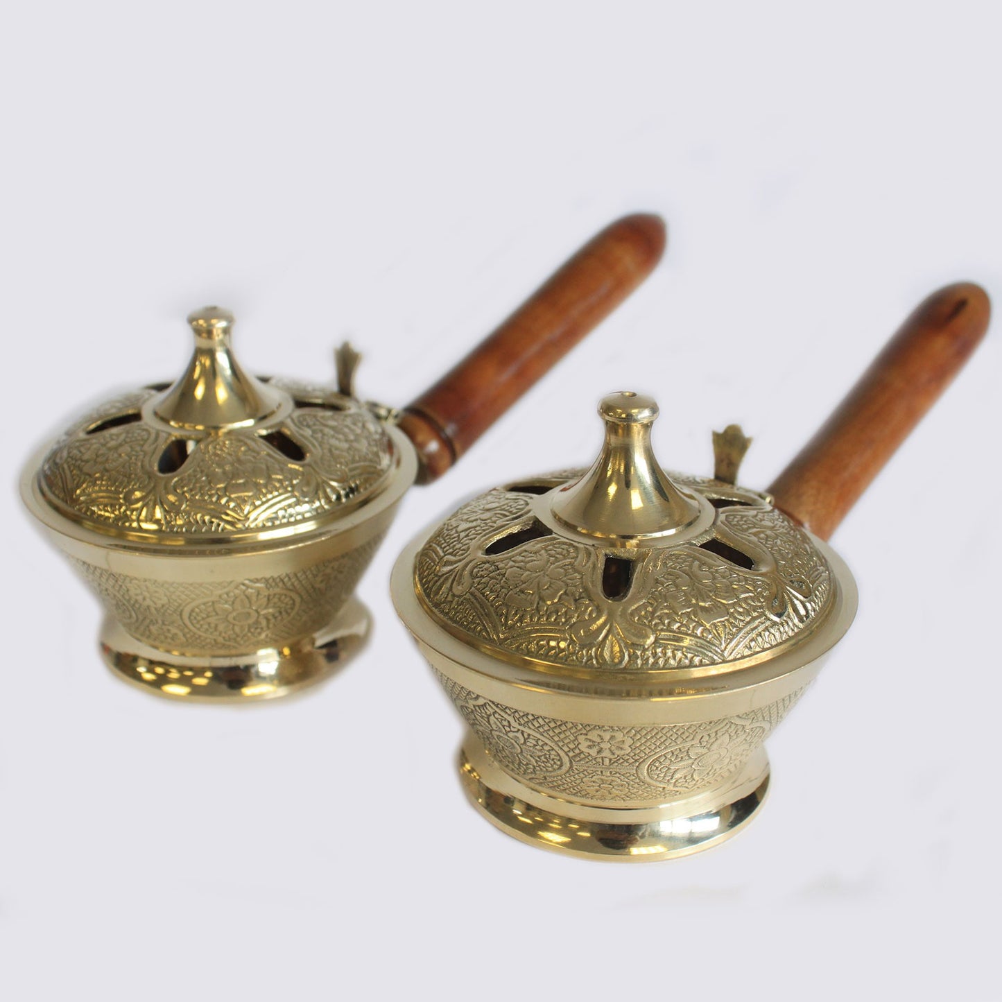 Two brass incense burners with ornate designs sit side by side, featuring wooden handles. They have intricate patterns and are placed against a plain, light-colored background.