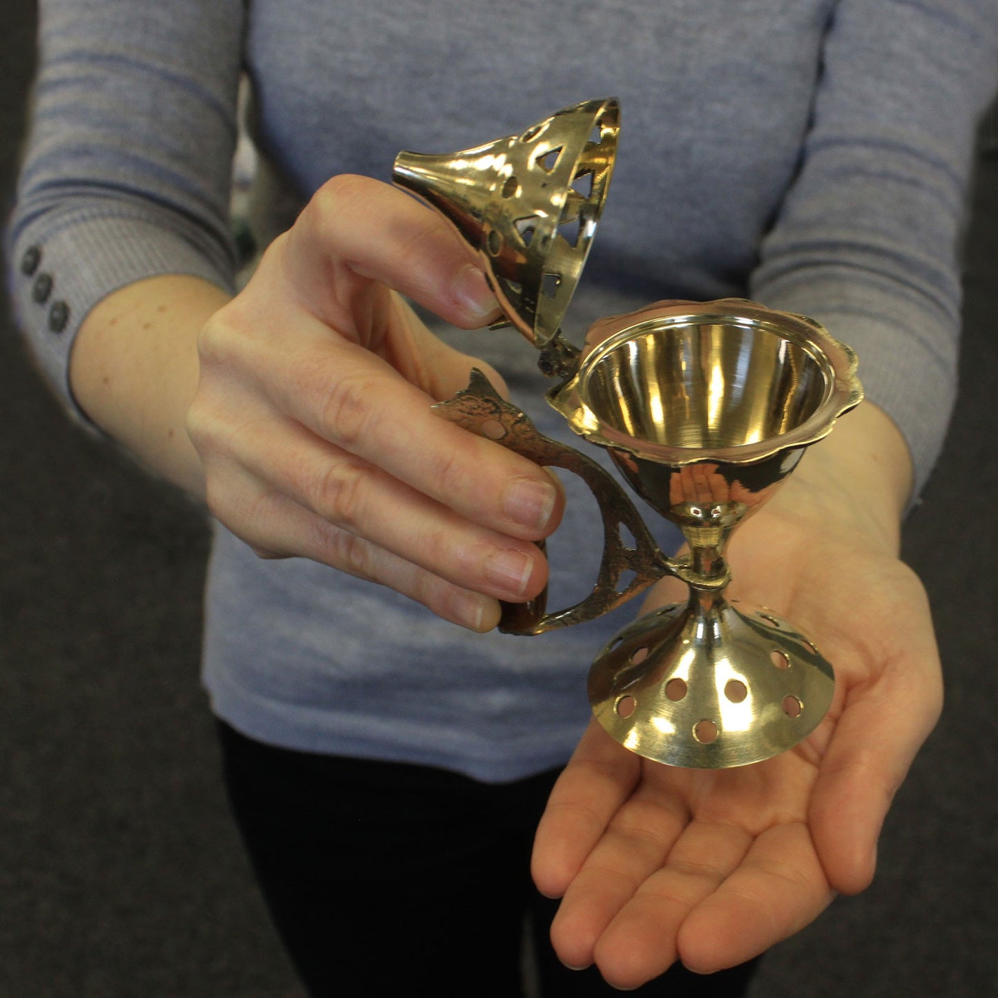 A person holds an ornate, perforated brass strainer with a hinged lid open, displayed against a blurred indoor background.