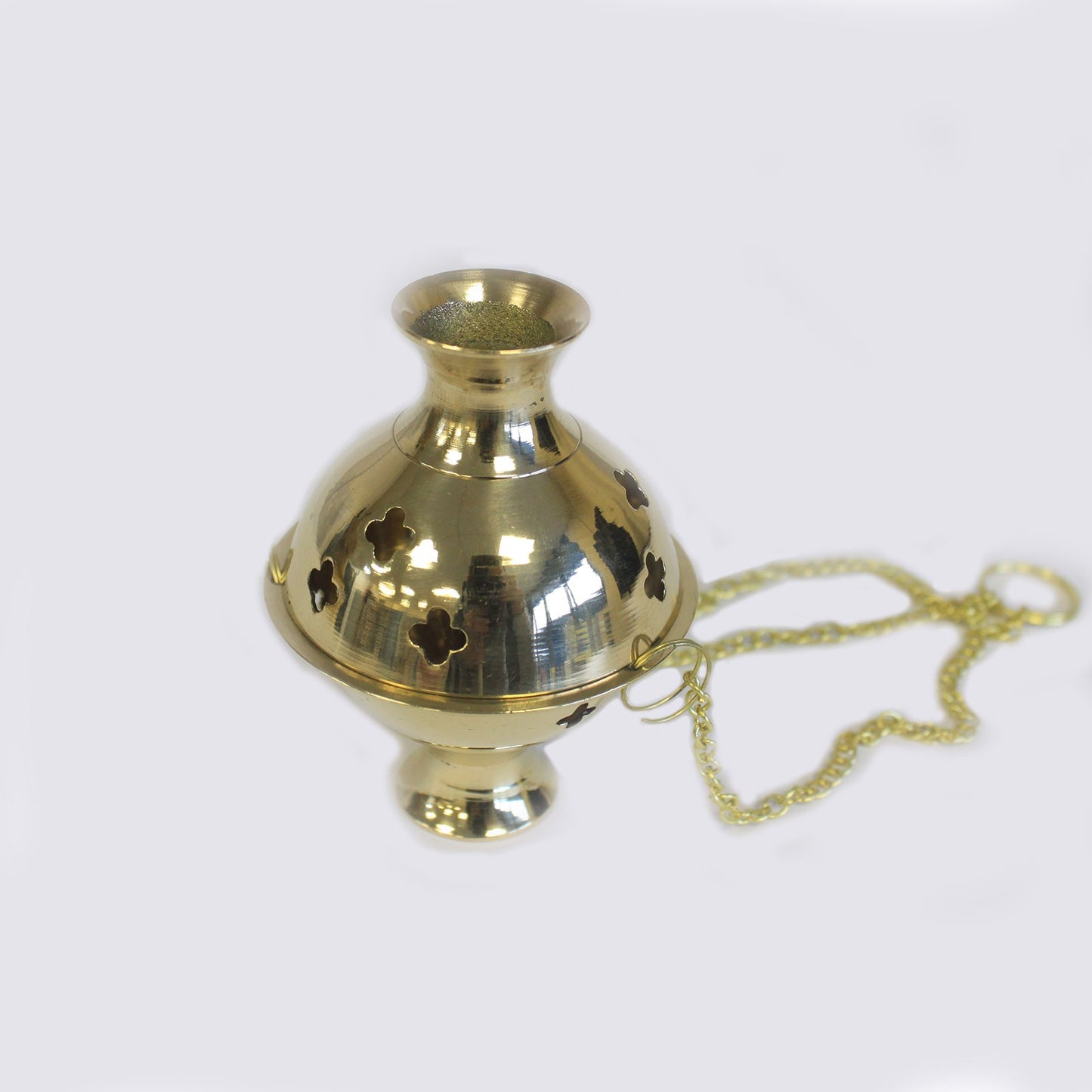 A polished brass censer with clover-shaped cutouts is connected to a chain, resting against a plain, light background.