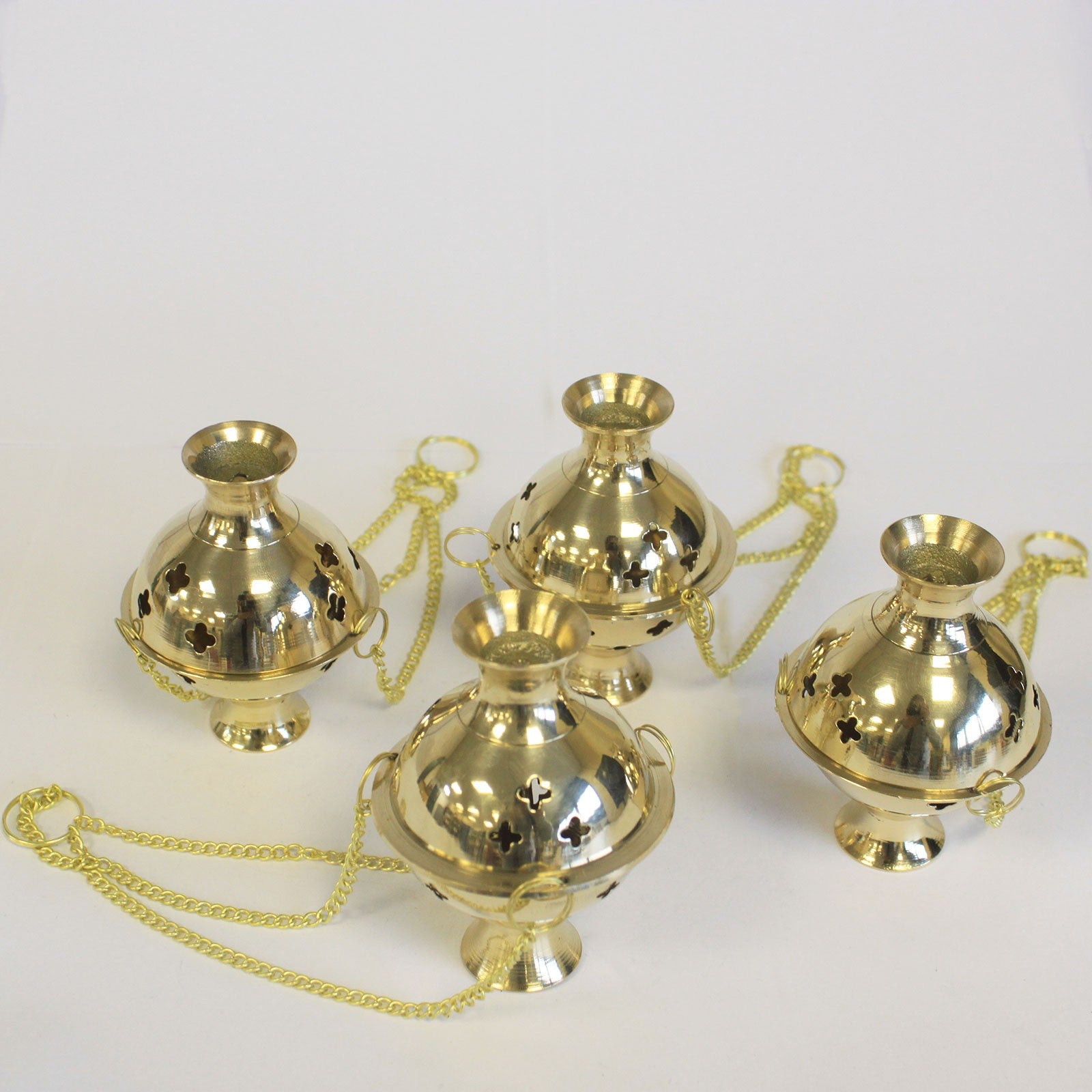 Four polished brass incense burners with decorative cutouts and chains are placed on a white surface, arranged in a close cluster, reflecting light in an indoor setting.