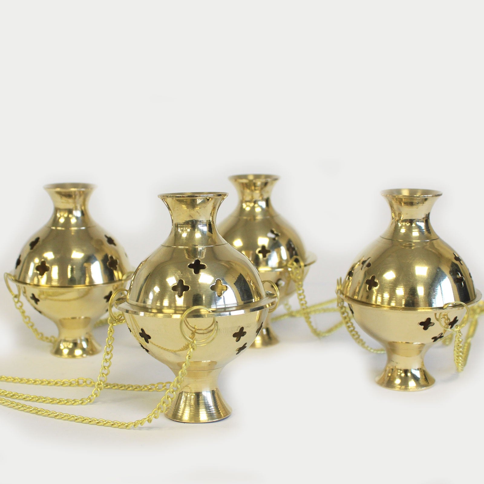 Four shiny brass incense burners with intricate cut-out designs rest on a white surface, surrounded by linked chains for holding or carrying.