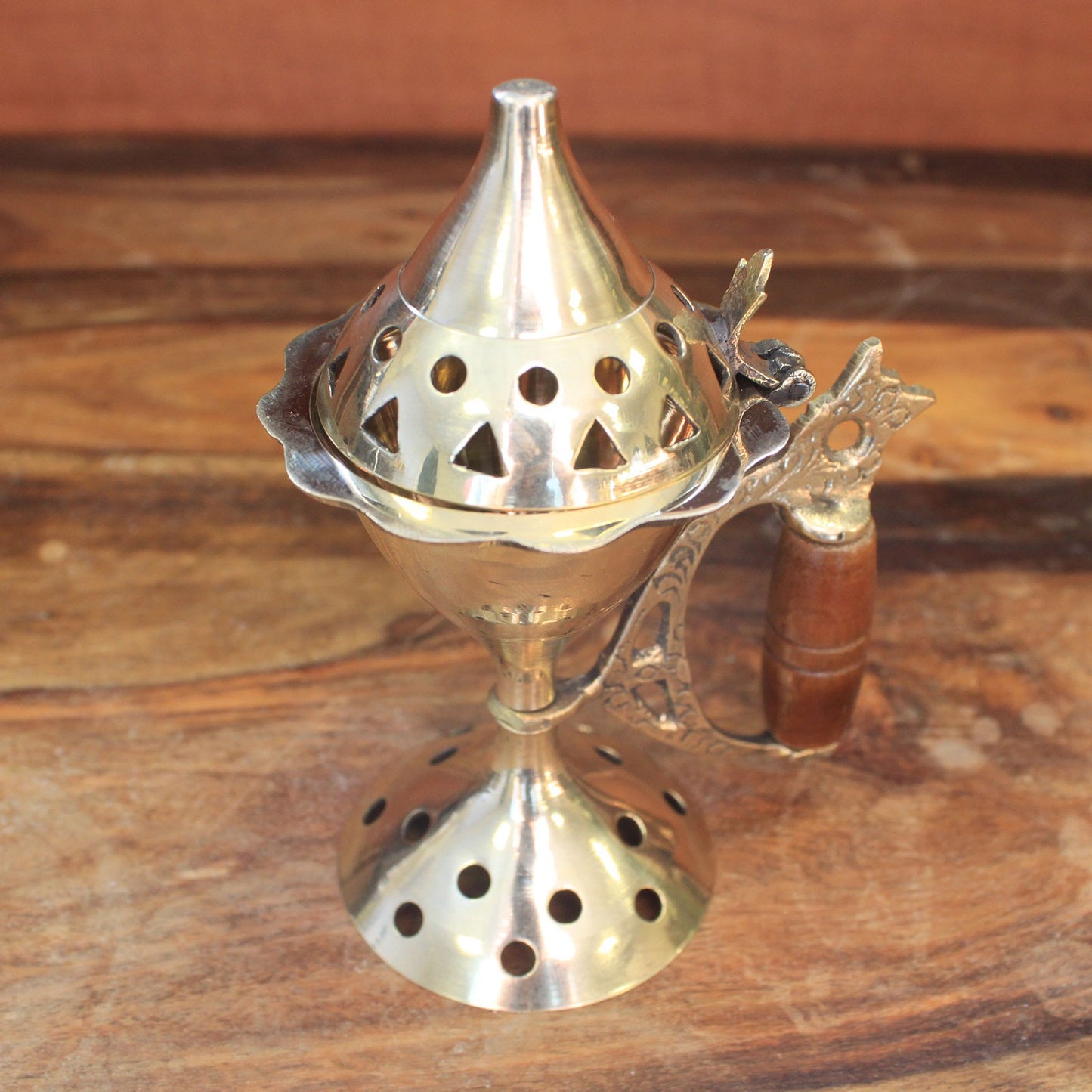 A brass incense burner with a conical, perforated lid rests on a wooden table. It features a decorative handle and is designed to release fragrant smoke through its geometric openings.