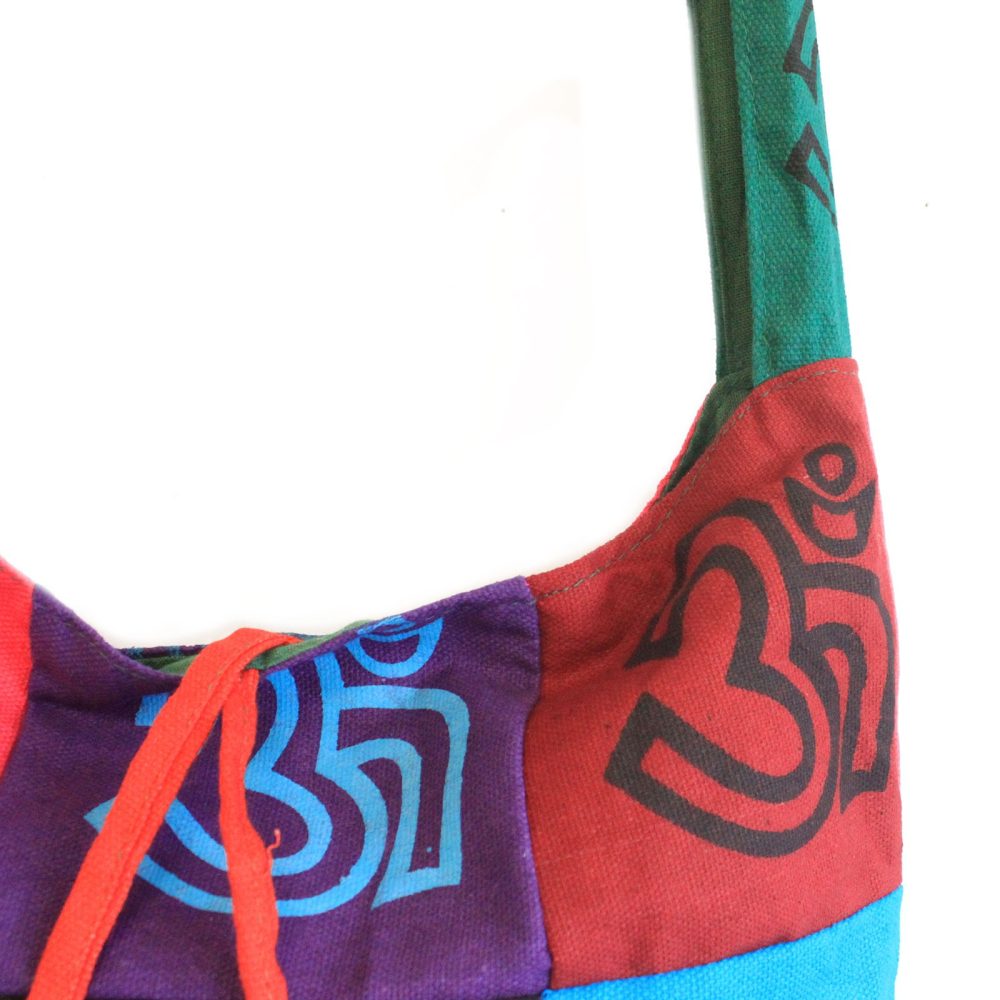 A patchwork bag with a shoulder strap displays Om symbols, tied with orange cords. Colors include purple, red, green, and blue. The bag is against a white background.