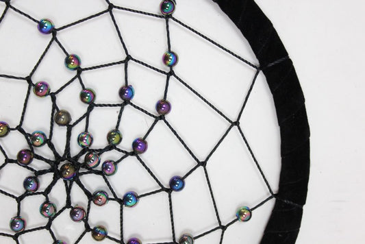 A dreamcatcher, adorned with iridescent beads, is strung with black thread onto a black velvet hoop; it is displayed against a white background.