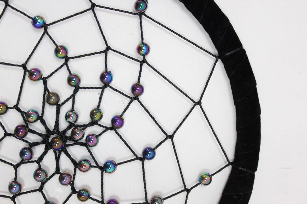 A dreamcatcher, adorned with iridescent beads, is strung with black thread onto a black velvet hoop; it rests against a white background.