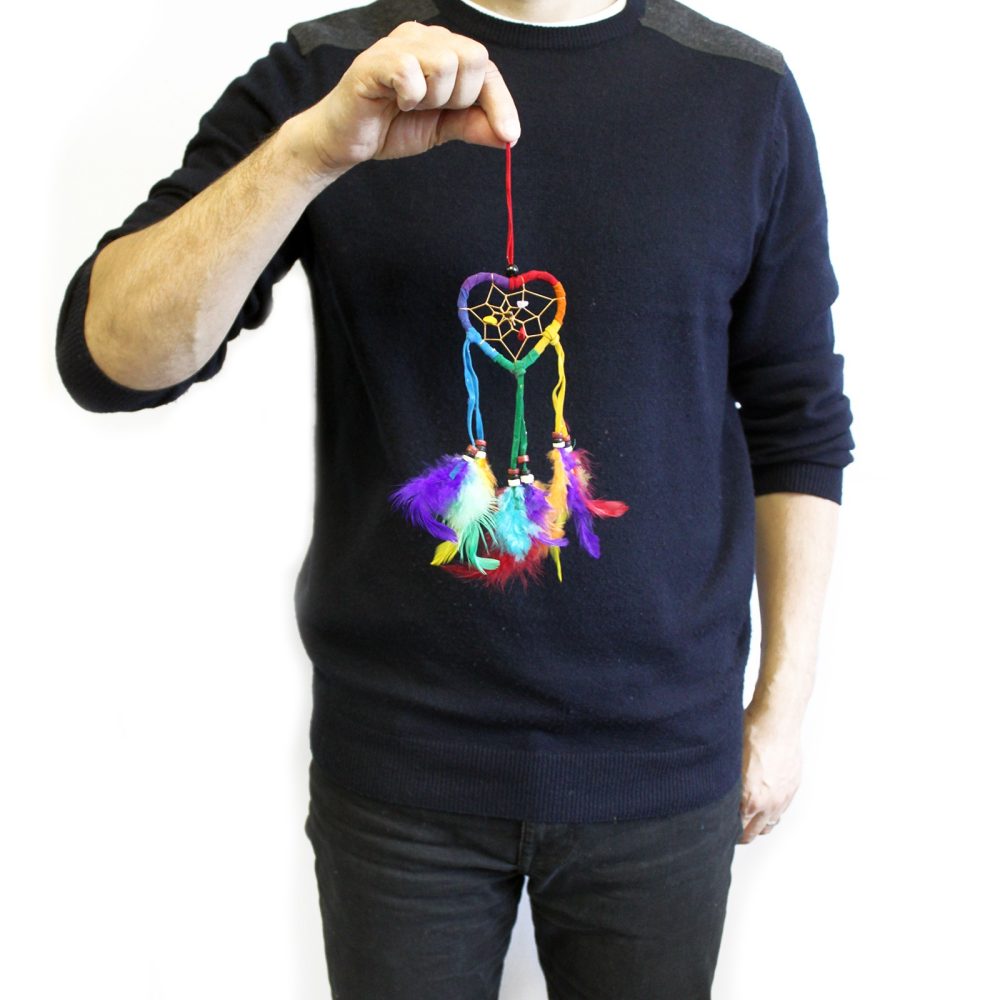 A multicolored heart-shaped dreamcatcher, adorned with feathers, is held by a person wearing a dark sweater and jeans against a white backdrop.