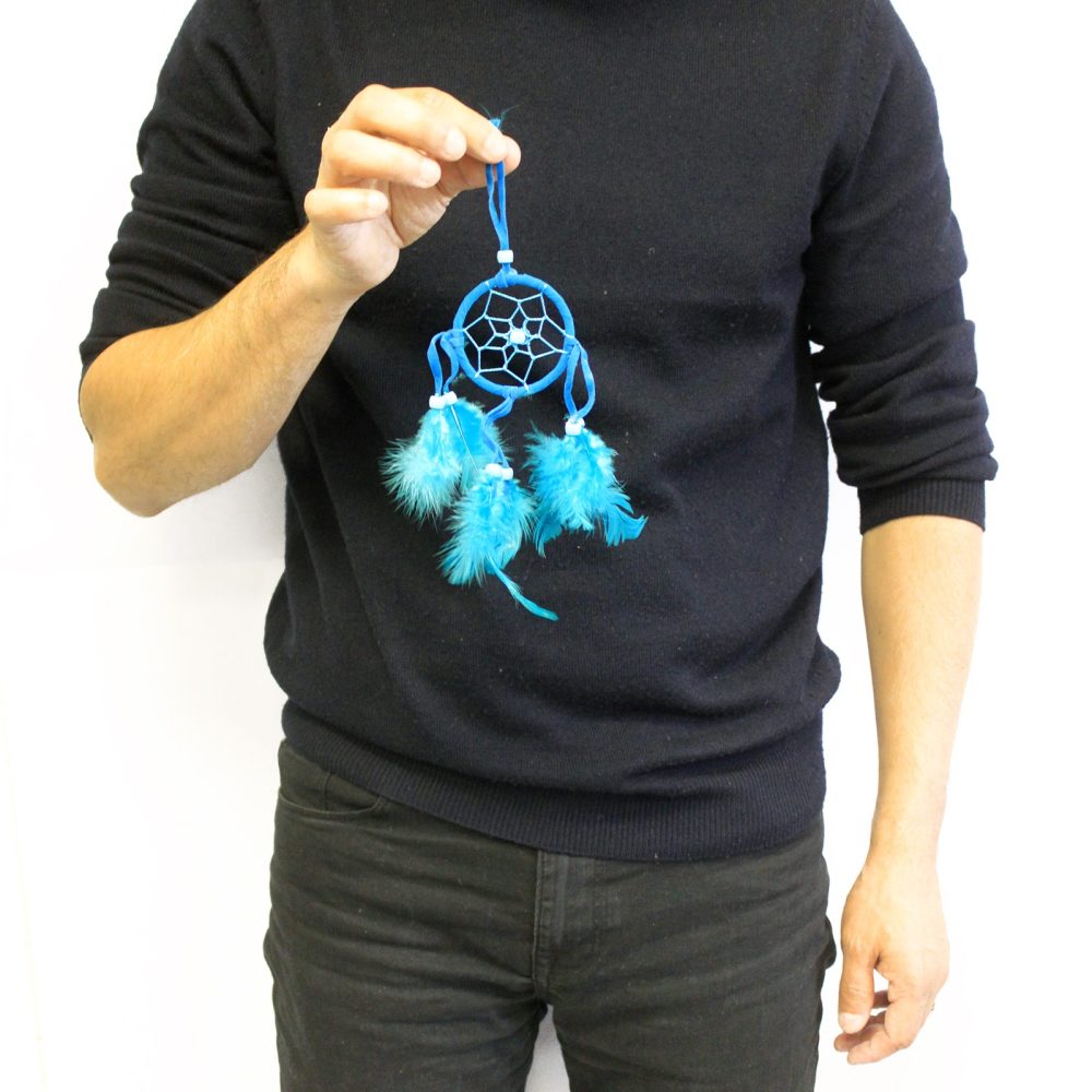 A person holds a small, blue dreamcatcher adorned with feathers. The dreamcatcher has a circular net and three blue feathers. The person is wearing a black sweater and dark pants, standing against a white background.