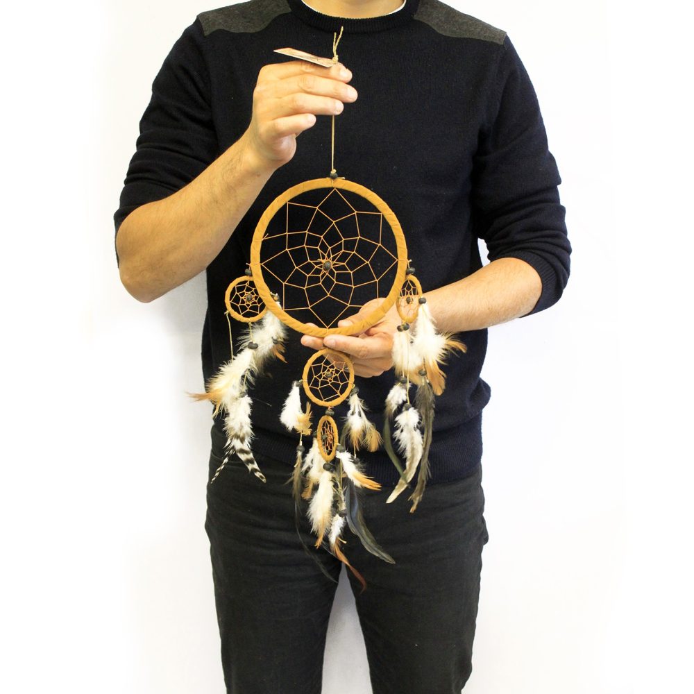 A person holds a tan dreamcatcher with feathers, featuring three smaller rings. The dreamcatcher is suspended by a string. The person is wearing a dark sweater and dark pants, against a white background.