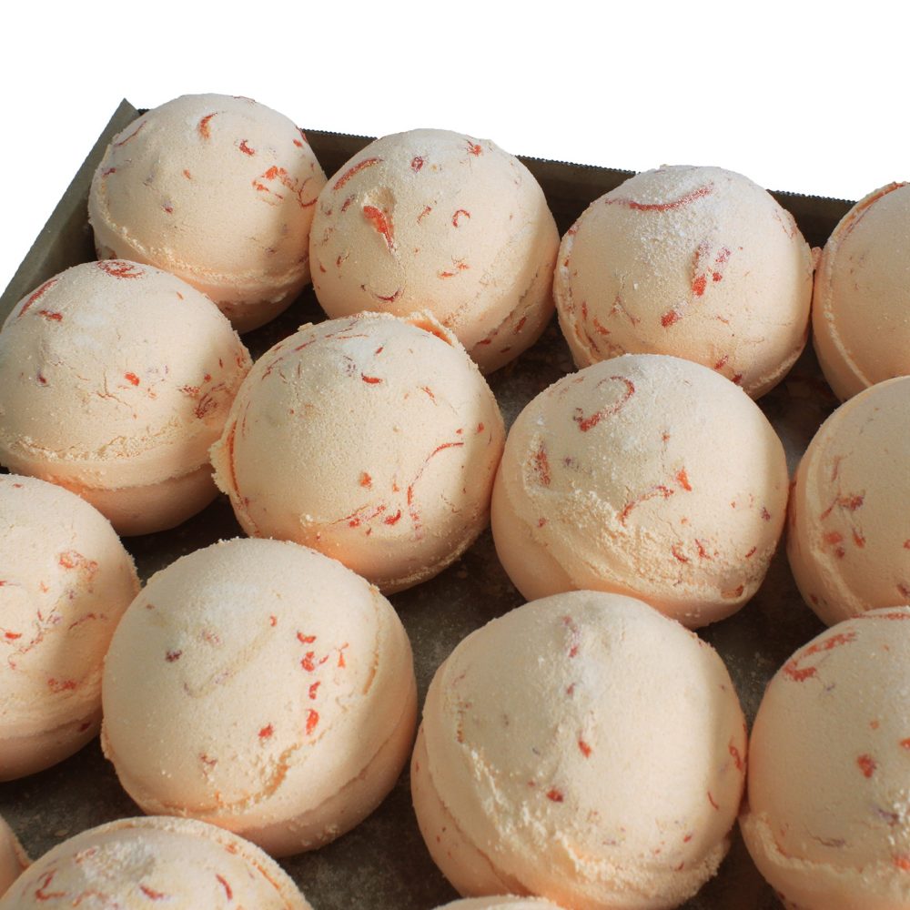 Round, orange bath bombs with red speckles sit arranged in rows on a tray, showcasing a uniform, textured surface in a neatly organized context.