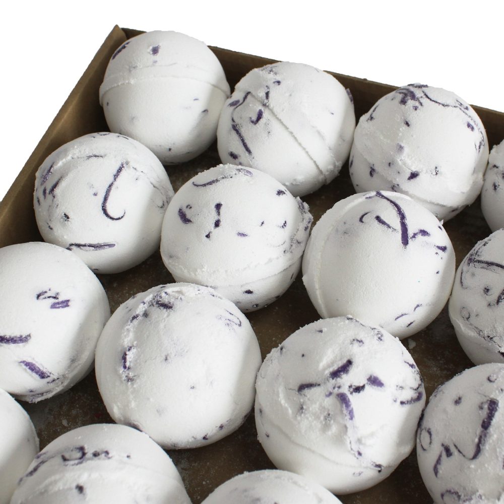White bath bombs speckled with purple flecks are displayed in a cardboard box, arranged in neat rows.