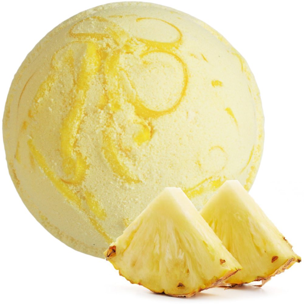 A round, yellow bath bomb with swirling patterns is accompanied by two slices of fresh pineapple in a bright setting.