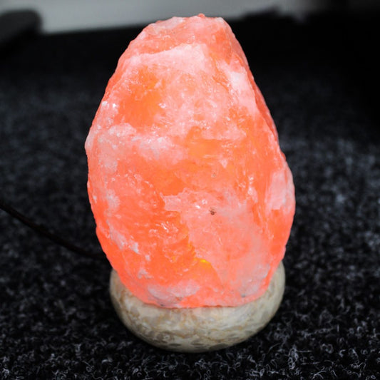 A glowing pink Himalayan salt lamp, emitting warm light, sits on a textured grey surface with a black cord extending from its base.