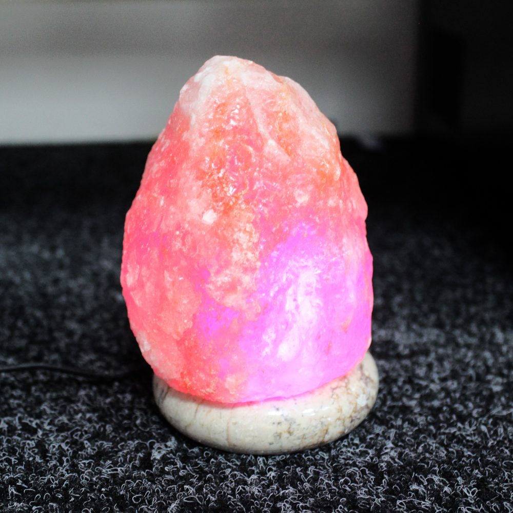 A pink Himalayan salt lamp glows softly, emitting a warm light on a textured gray carpet. The lamp is mounted on a smooth, round base.