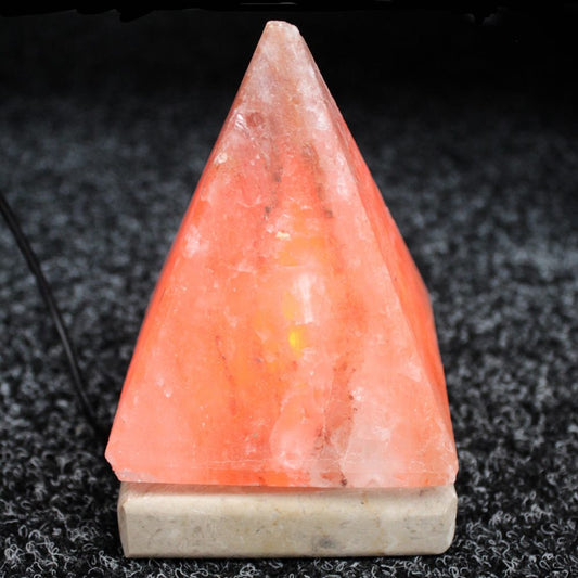 A pink Himalayan salt lamp shaped like a pyramid glows warmly from within, sitting on a square base. It's placed on a textured dark surface, with a power cord visible.
