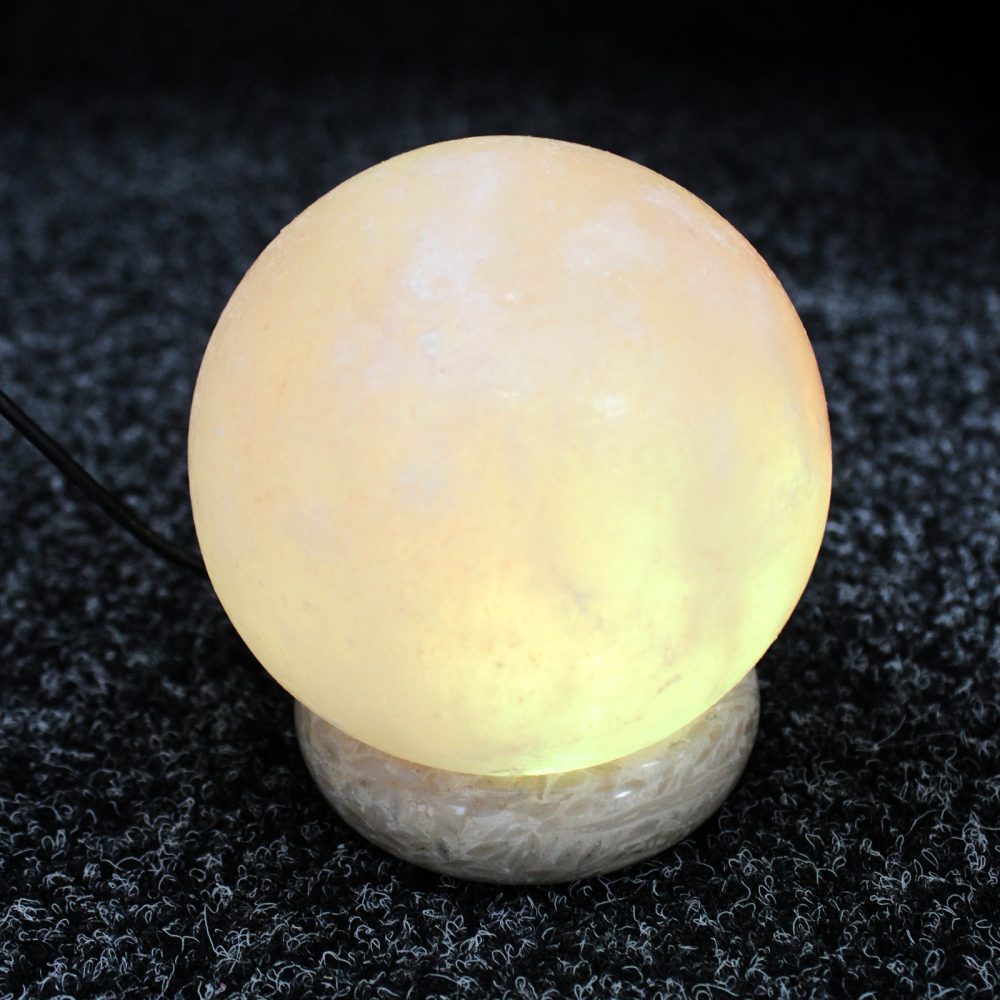 A glowing spherical lamp rests on a textured dark surface, emitting a warm yellow light and is connected by a visible black cord.