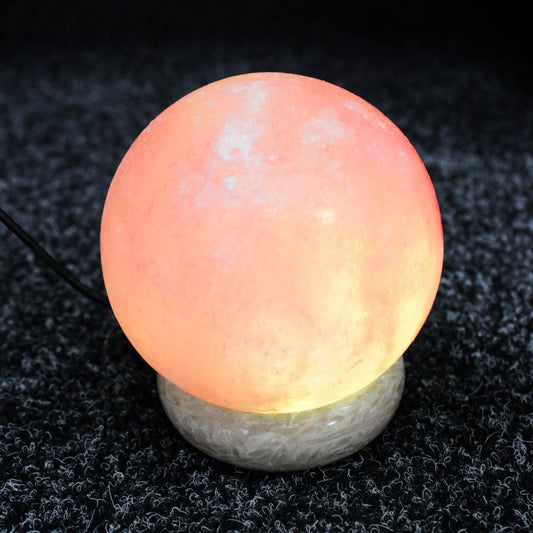 A spherical salt lamp emits a warm, orange glow atop a textured, circular base, situated on a dark, carpeted surface.