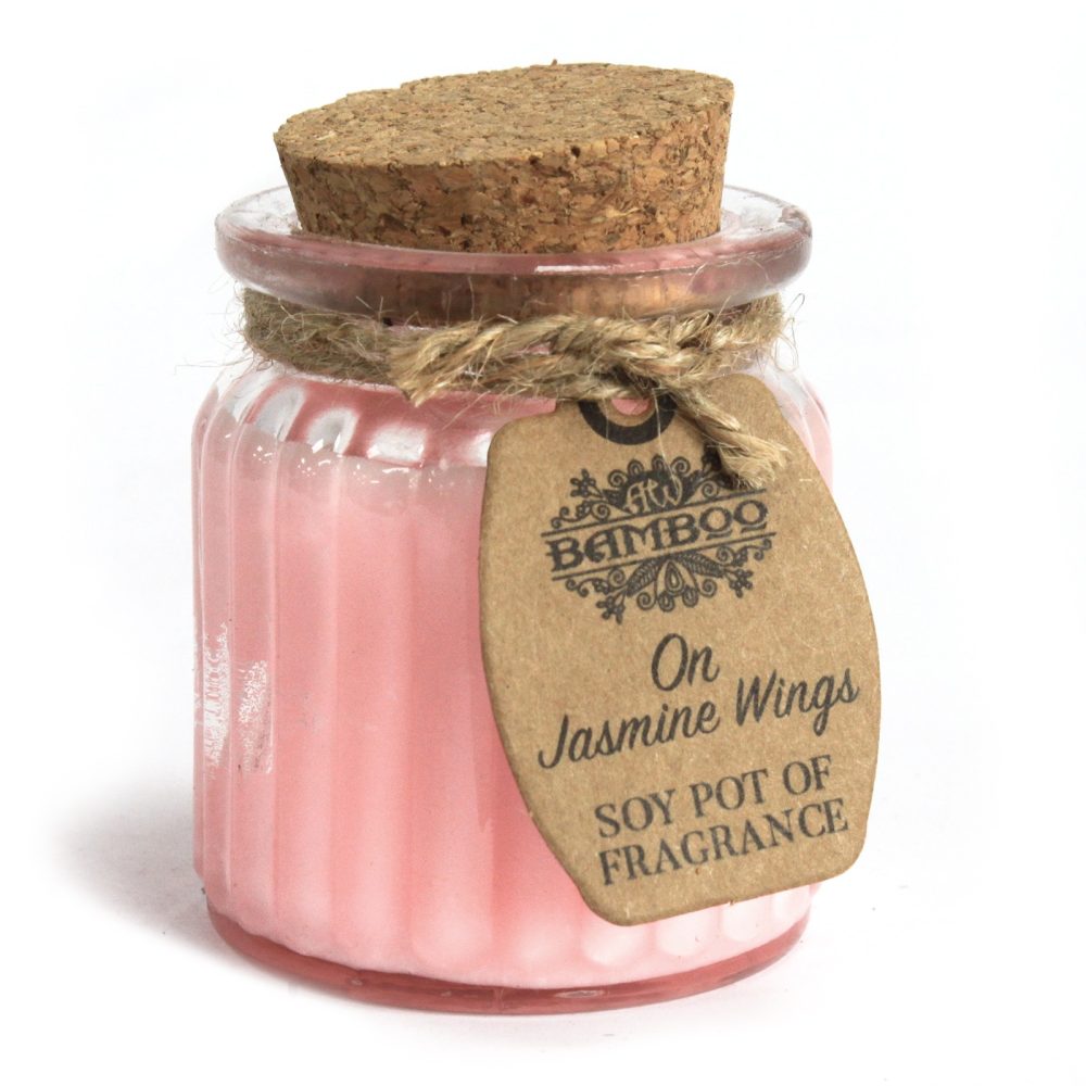 A pink soy candle sits on a white surface, housed in a ribbed glass jar with a cork lid. A tag reads "BAMBOO On Jasmine Wings SOY POT OF FRAGRANCE."