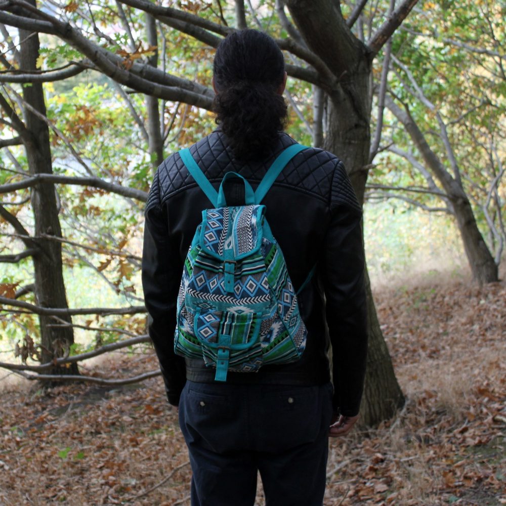 A person with long dark hair faces away from the camera, wearing a black jacket and turquoise backpack. They stand on a leaf-covered path among trees in a wooded area.