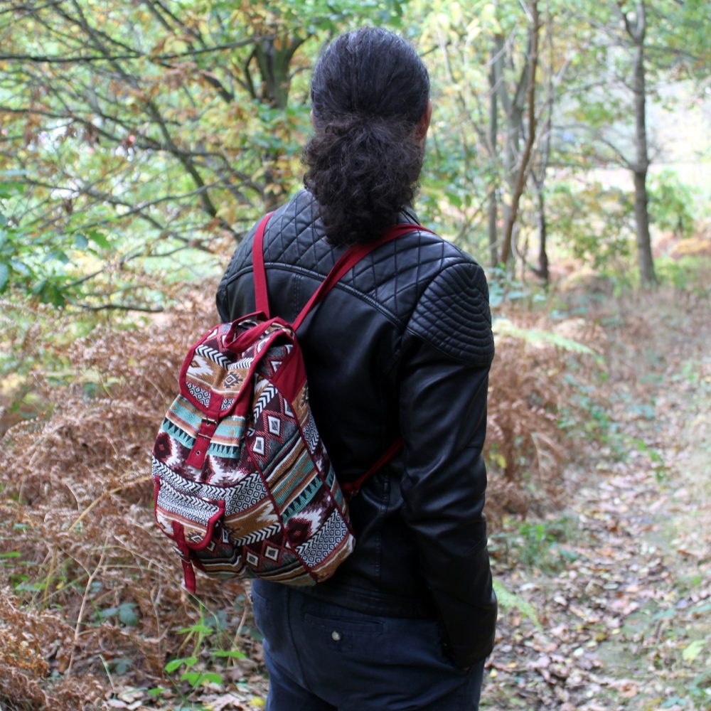 A person, wearing a black leather jacket and printed backpack, walks away from the camera along a trail in a wooded area.