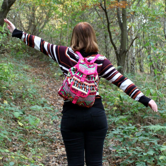 A woman with a colorful backpack walks away down a leaf-strewn path, arms outstretched. She's wearing a striped sweater and dark pants in a wooded area.