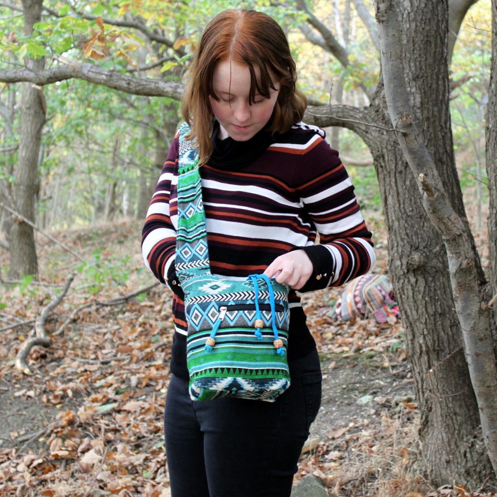 A young woman looks into a blue and green patterned shoulder bag, standing in a wooded area with fallen leaves on the ground.