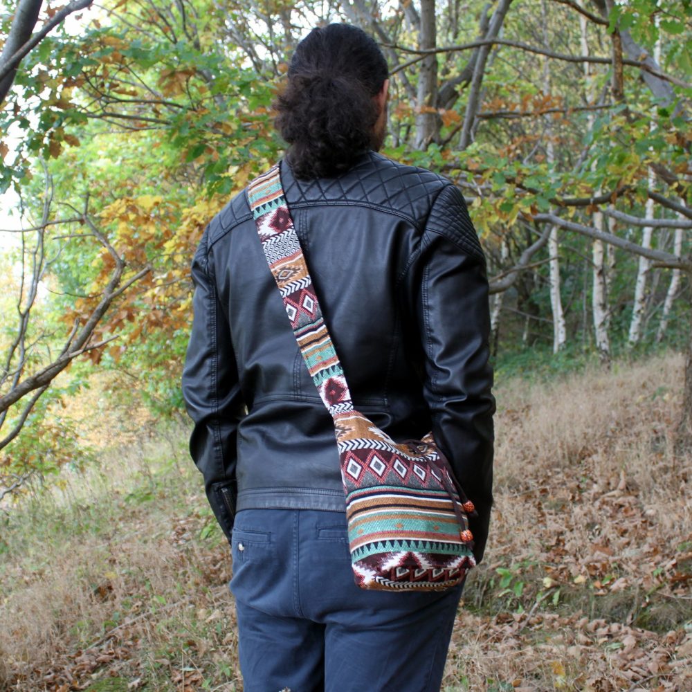 A person with long, curly hair wears a decorative, patterned bag over their shoulder. They wear a black leather jacket and blue pants, and are standing in a wooded, outdoor setting.