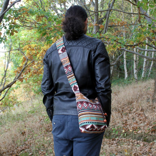 A person with long, curly hair wears a decorative, patterned bag over their shoulder. They wear a black leather jacket and blue pants, and are standing in a wooded, outdoor setting.