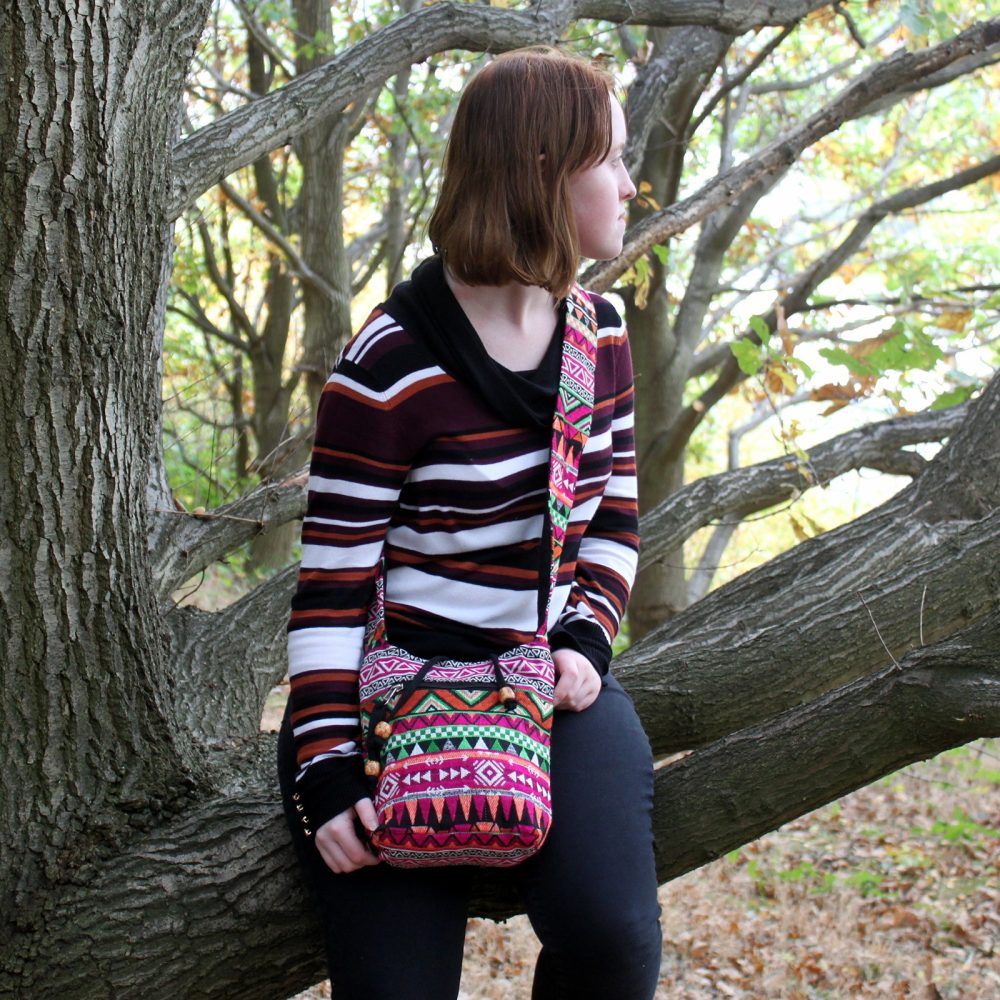 A young woman perches on a tree branch, gazing right. She wears a striped sweater, black pants, and a colorful, patterned crossbody bag in a leafy outdoor setting.
