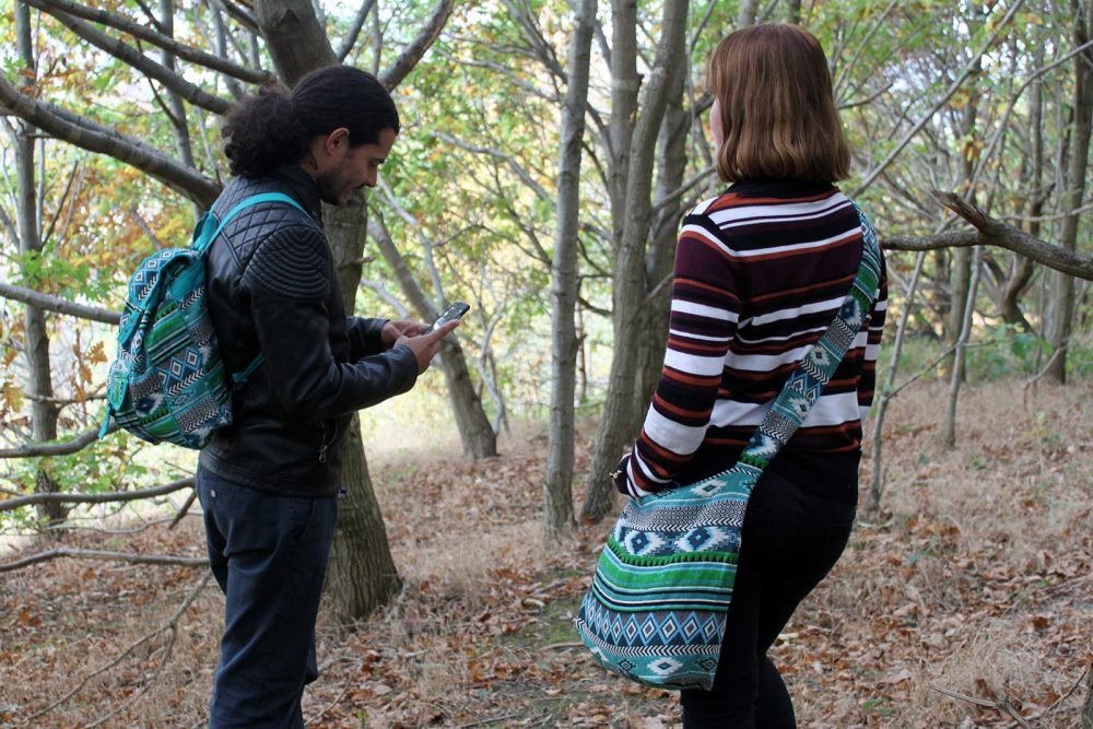 A man is looking at his phone in the woods while wearing a backpack, and a woman stands next to him with a bag.