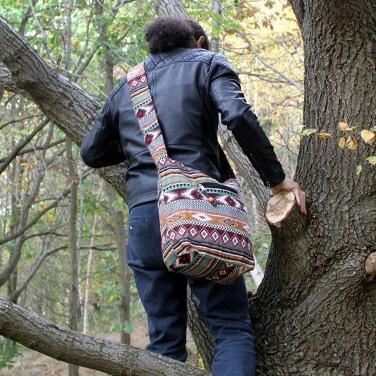 A person with a colorful shoulder bag is climbing a tree in a forest. They are wearing a black leather jacket and blue pants.