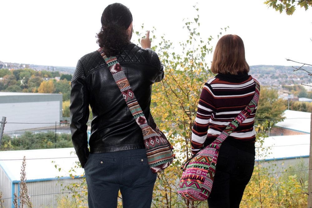 Two people, back view, stand overlooking a city on a hill. Both have patterned shoulder bags. One points into the distance.
