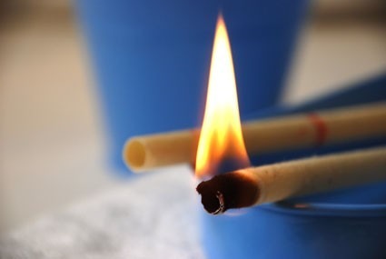 A thin rolled stick burns at its charred tip while a second lies beside it, flame glowing above a blue bowl on a soft white cloth with blurred blue background.