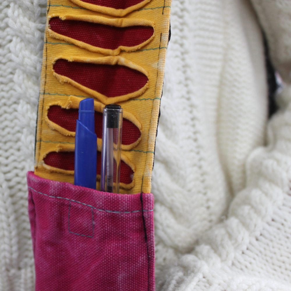 Two pens sit in a pink pocket, which is part of a yellow strap with red cutouts. The strap is against a textured white sweater.