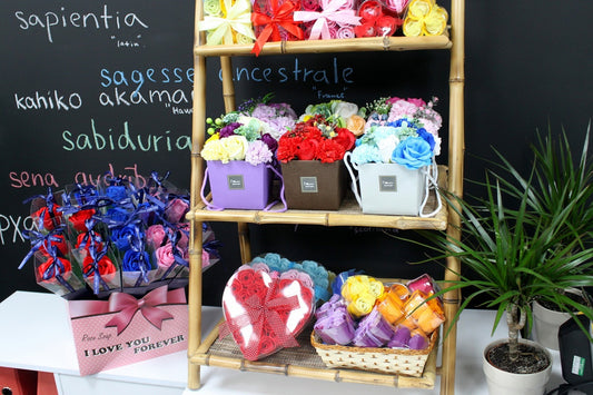 Colorful artificial flower arrangements sit on a bamboo shelf and table in front of a black chalkboard with handwritten words; visible text: sapientia "latin", sagesse ancestrale "France", kahiko akamai "Hawa", sabiduria, Rose Soap, I LOVE YOU FOREVER.