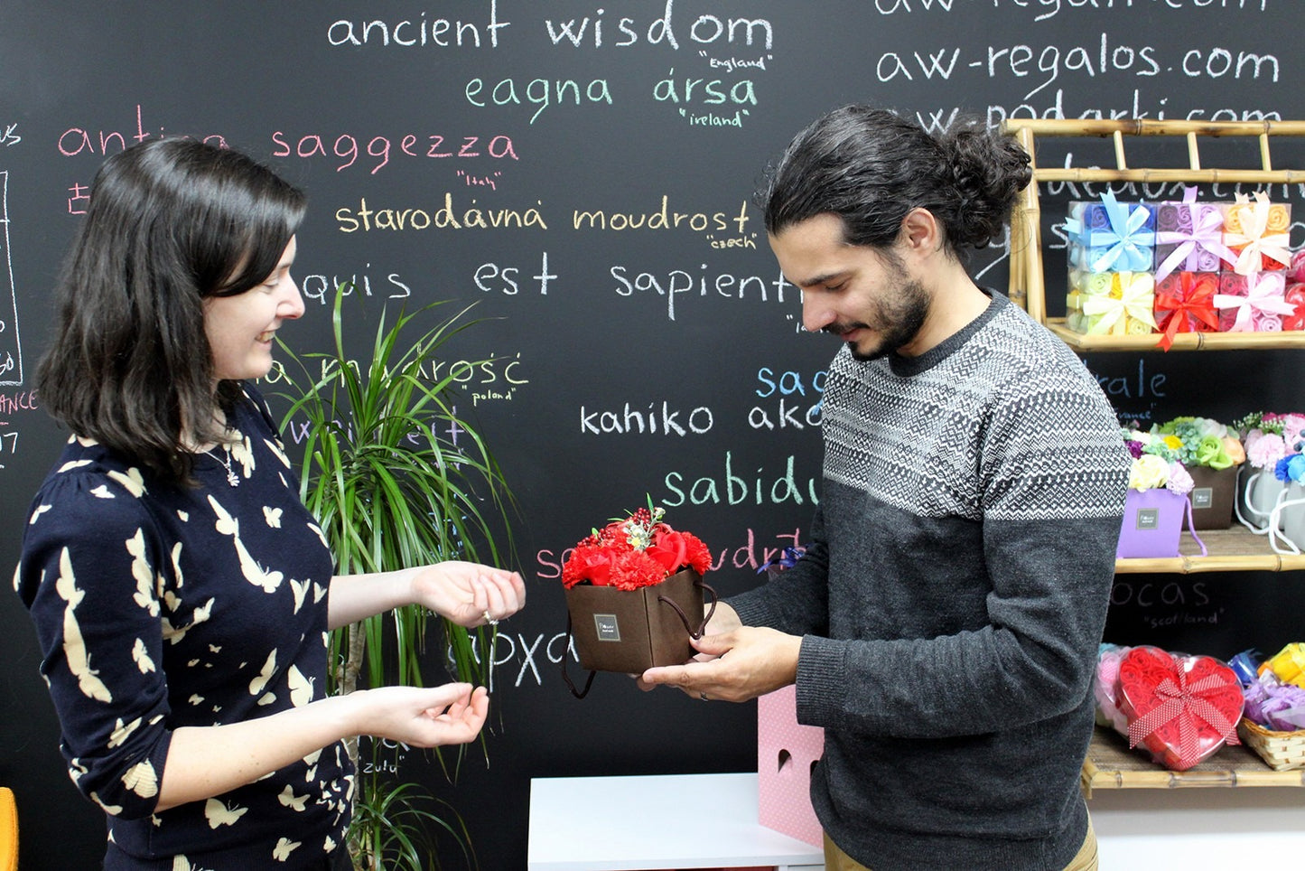 A man hands a box of red flowers to a smiling woman in front of a blackboard with the words “ancient wisdom eagna ársa saggezza starodávna moudrost quis est sapiens kahiko ako sabidi aw-regalos.com w-podarki.com”.